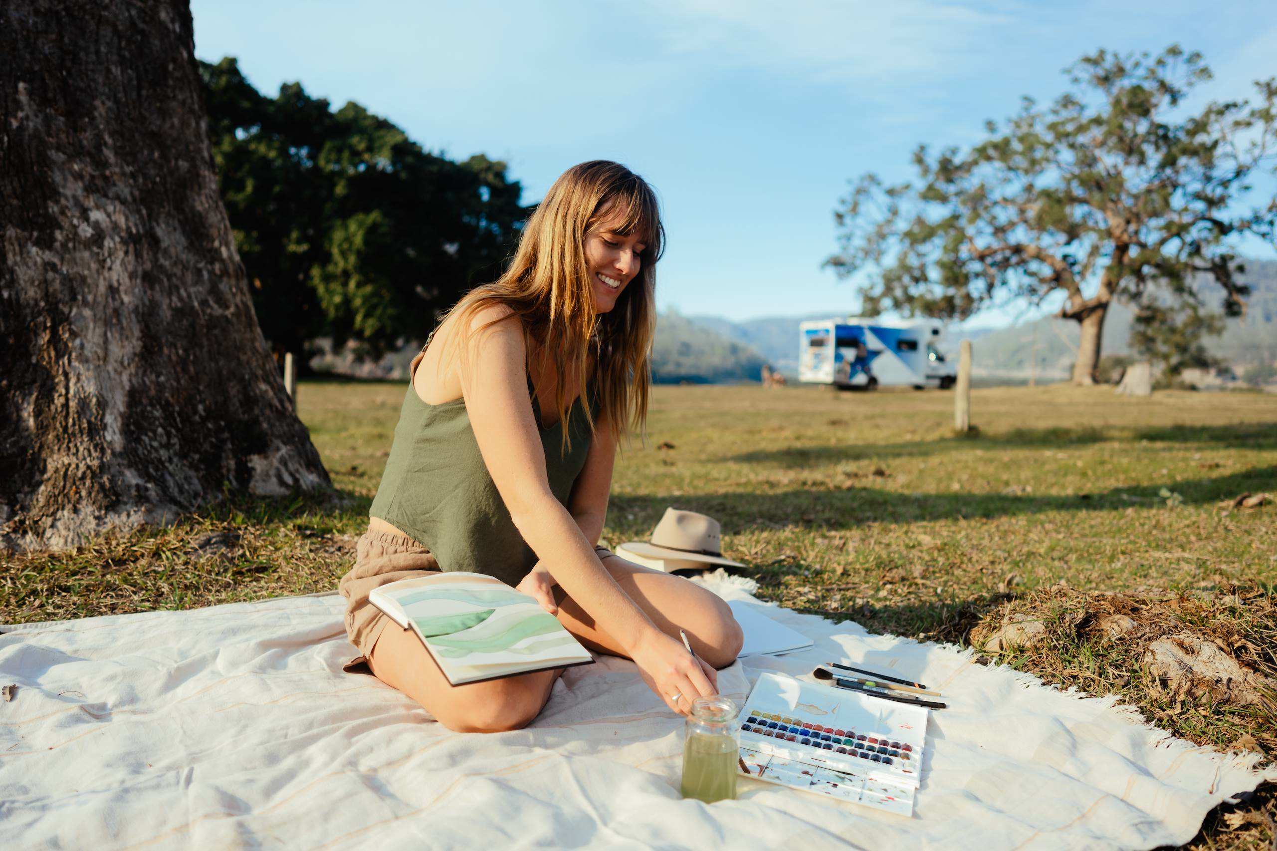 Lady painting on picnic blanket under a tree with Star RV polaris-6 parked behind