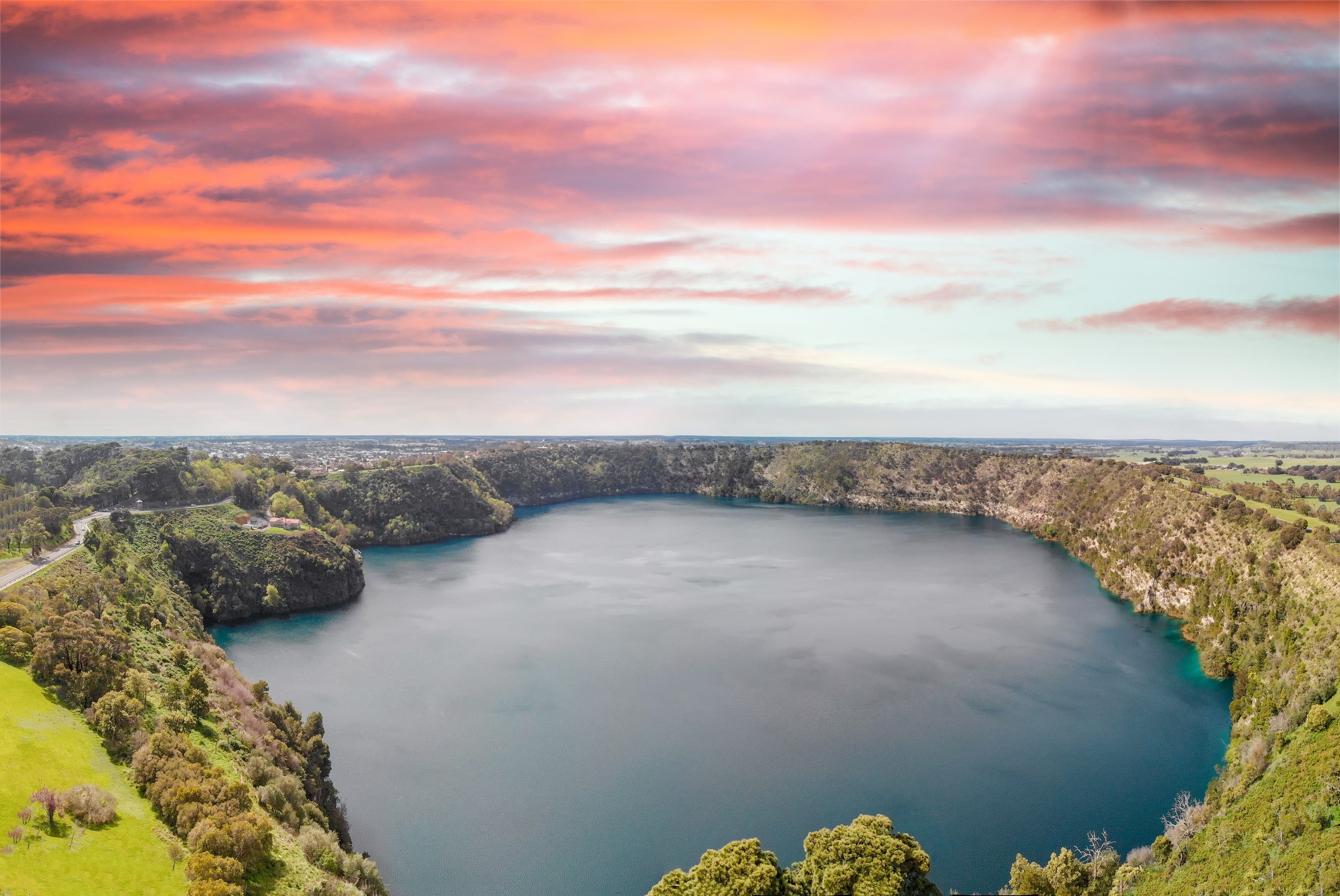 A stunning aerial view of Blue Lake in Mount Gambier, showcasing the vibrant blue water surrounded by lush greenery under a colorful sunset sky.
