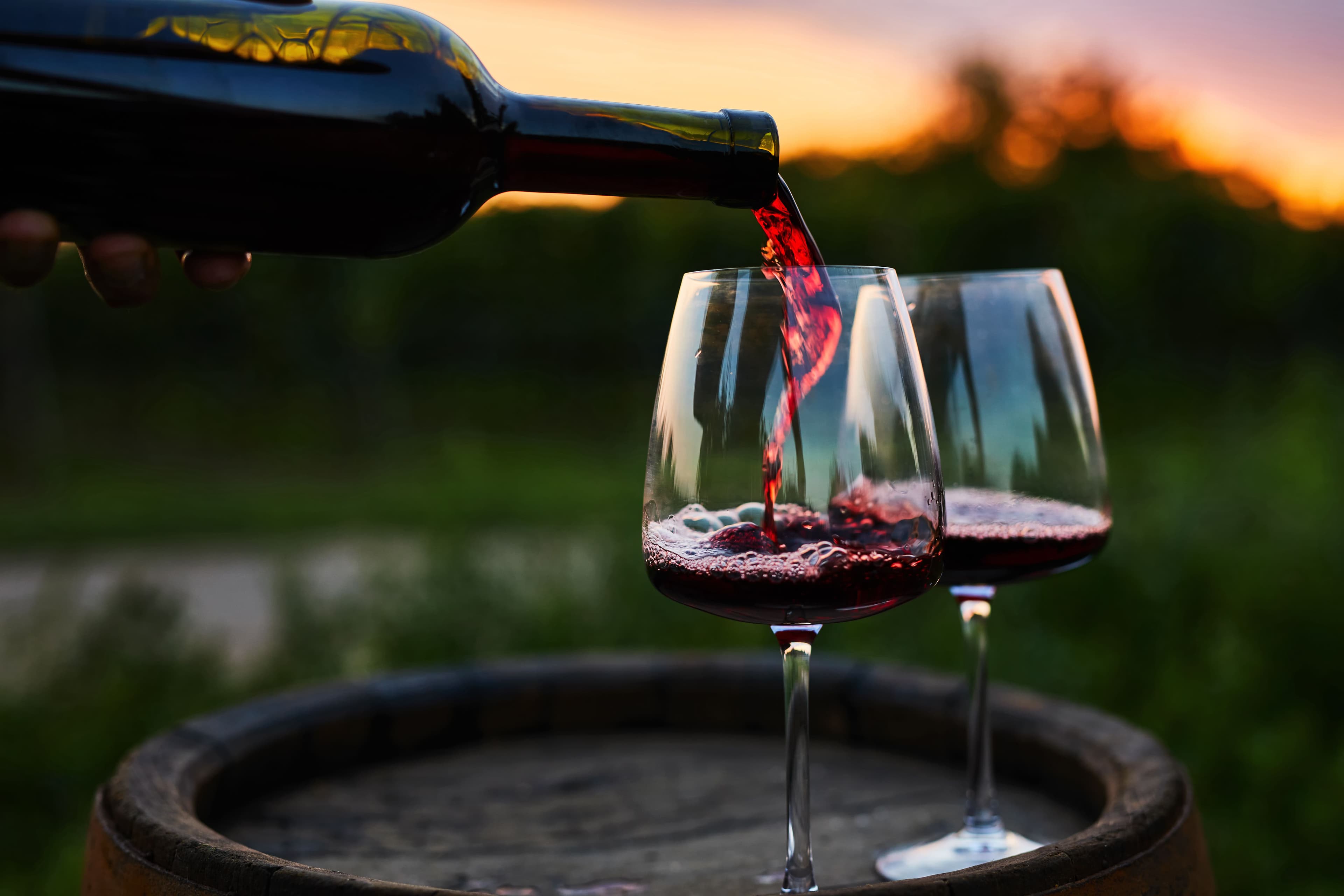 An inviting scene of red wine being poured into two glasses set on a wine barrel, with a picturesque Coonawarra vineyard sunset in the background.