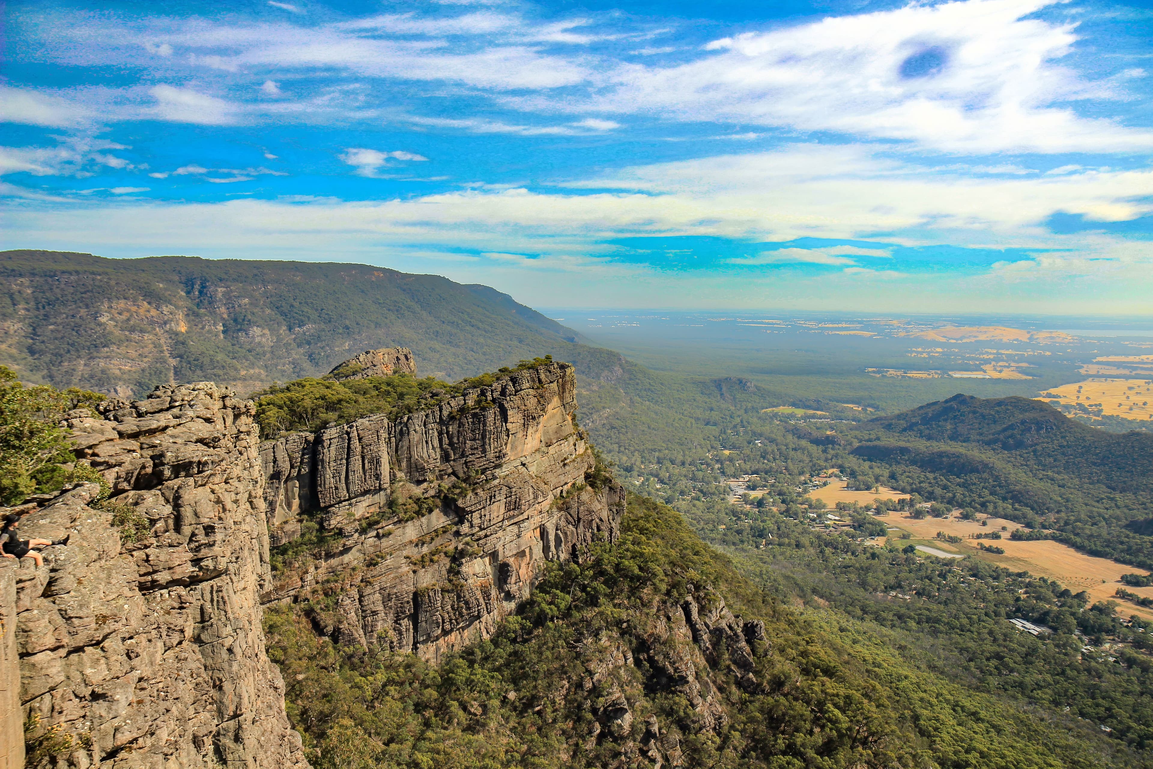 A breathtaking view of the rugged landscape of Grampians National Park, featuring dramatic sandstone peaks, dense forests, and cascading waterfalls under a clear blue sky.