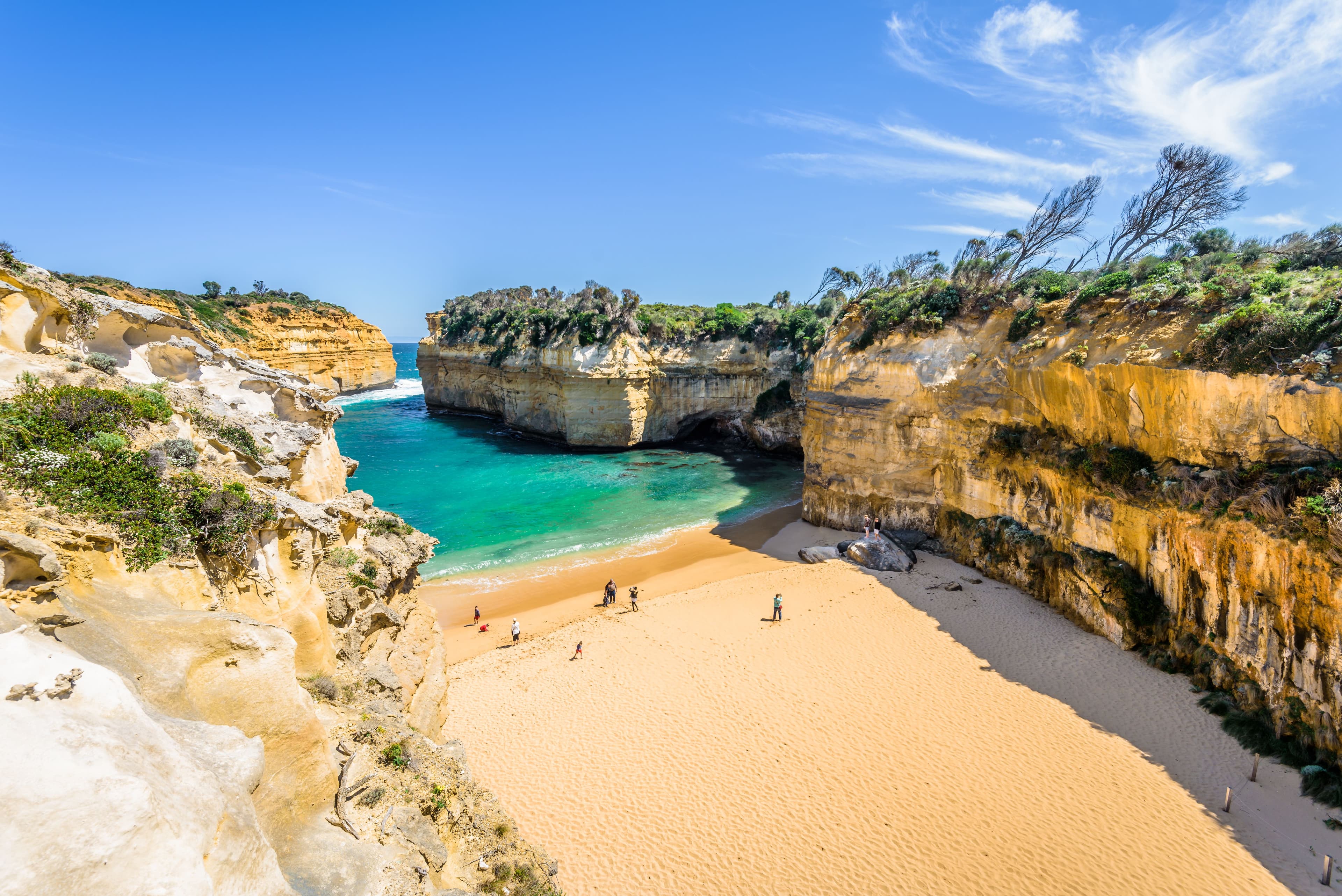 A striking coastal scene of Loch Ard Gorge, showcasing towering limestone cliffs, pristine sandy beaches, and the turquoise waters of the Southern Ocean under a bright, sunny sky.